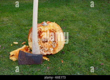 Axe stands against a large orange pumpkin, roughly cut in half outdoors, spilling flesh and seeds - with copy space Stock Photo