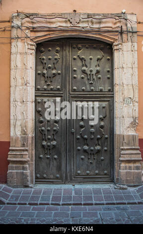 An old elaborate door, with beautifully crafted details and a massive stone frame, and a brick paved walkway, in Guanajuato, Mexico Stock Photo