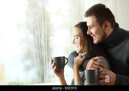 Side view portrait of a happy couple holding coffee mugs looking outside through a window a rainy day of winter at home Stock Photo