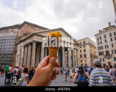 Hand holding ice cream in Rome. Ice cream cone with chocolate and mango flavor before blurred background of Pantheon landmark in Piazza della Rotonda. Stock Photo