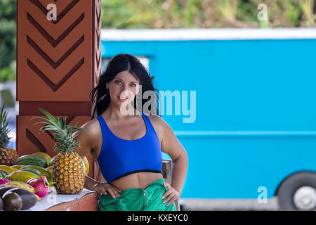 Slender healthy young woman with a fresh tropical pineapple posing leaning on a wall on an open air verandah with a blue truck and copy space behind , Stock Photo