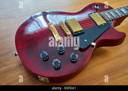 Shiny Wine Red Guitar With Golden Hardware Placed On A Wooden Table Angled Side View Focus On Stop Bar And Pickups Stock Photo