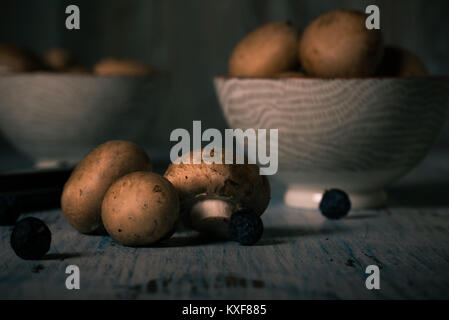 Horizontal photo of several edible mushrooms. Champignons spilled on vintage wooden board and others placed in two bowls. Dry blue berries spilled aro Stock Photo