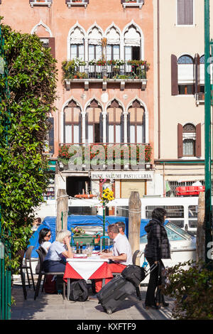 Tourists dining at open air restaurant tables in Cannaregio, Venice, Italy alongside the Cannaregio Canal viewed through greenery in the public park Stock Photo