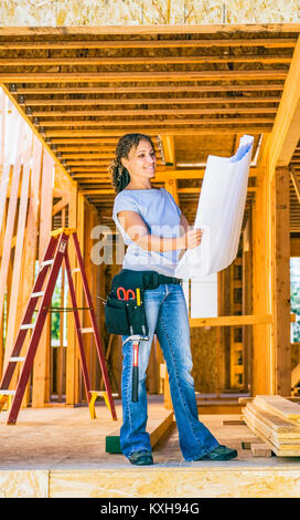 A early 30's mixed ethnicity woman wearing a tool belt and looking at construction plans in a home construction site  Model Release: 20060726 MR A Stock Photo