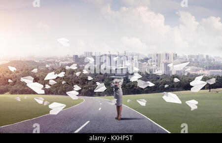 Business woman in suit with laptop instead of head keeping arms crossed while standing on the road among flying paper planes with beautiful landscape  Stock Photo