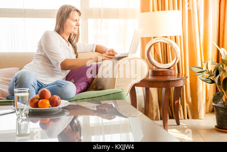 Mature attractive woman is working on laptop while sitting on a couch in her living room Stock Photo