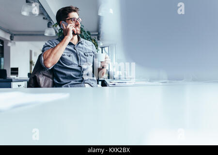 Young man with cup of coffee and talking on mobile phone. Male designer sitting in office having coffee and making phone call. Stock Photo