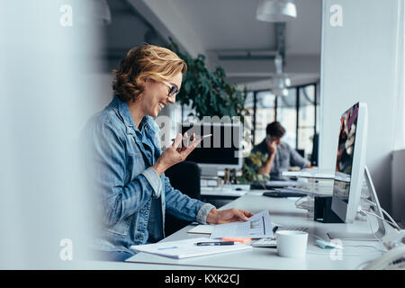 Businesswoman talking on smartphone and looking at documents. Young female executive sitting in front of pc. Stock Photo