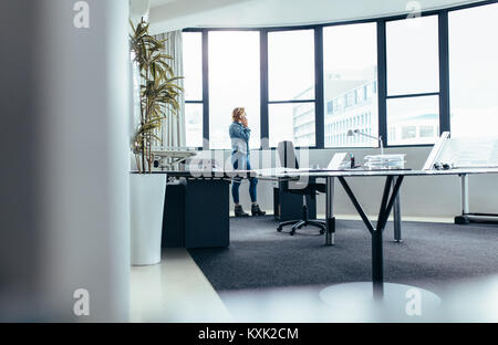 Young businesswoman standing in office and talking on mobile phone. Female manager standing by office window and using phone. Stock Photo