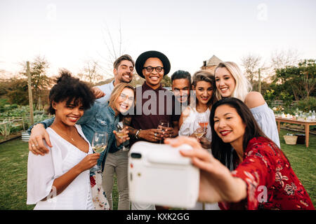 Woman with friends taking selfie using instant camera. Group selfie at outdoor party. Stock Photo