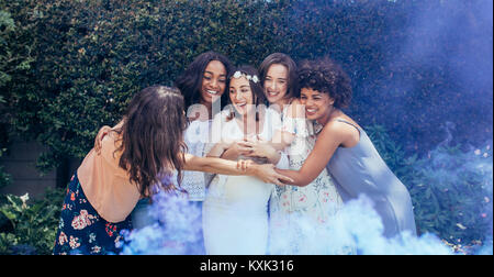 Group of smiling multi-ethnic female friends hugging pregnant woman outdoors. Expecting mother and her friends with smoke grenade at baby shower. Stock Photo