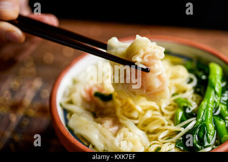 closeup of a young man getting some noodles with his chopsticks from an earthenware bowl with shrimp wonton noodle soup with choy sum, placed on a tab Stock Photo