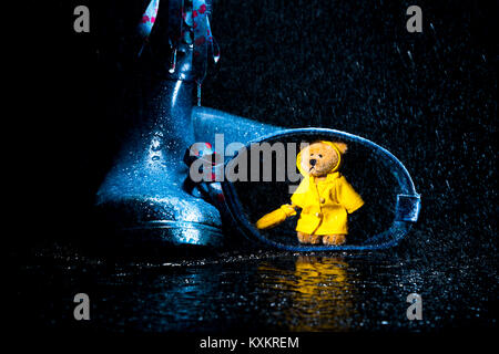 Little Teddy Bear in the yellow raincoat holding umbrella finding a refuge in a blue wellie. Stock Photo