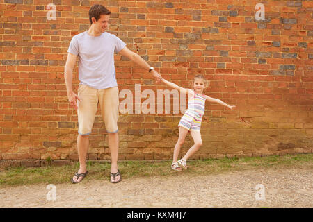 Girl leaning sideways holding father's hand by brick wall Stock Photo