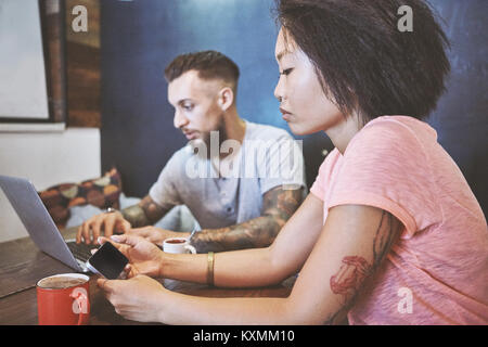 Multi ethnic hipster couple in cafe looking at smartphone and laptop,Shanghai French Concession,Shanghai,China Stock Photo