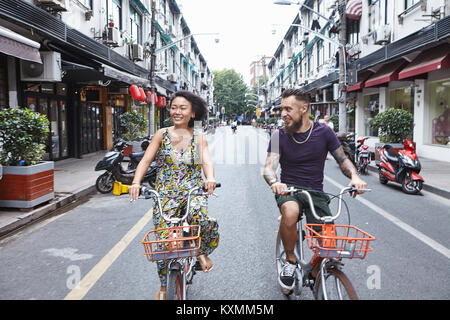 Multi ethnic hipster couple cycling along city street,Shanghai French Concession,Shanghai,China Stock Photo