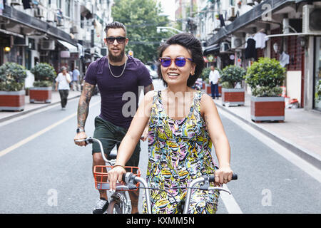 Multi ethnic hipster couple cycling along city street,Shanghai French Concession,Shanghai,China Stock Photo