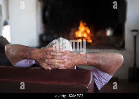 Rear view of senior man at home with hands behind his head in front of log fire Stock Photo