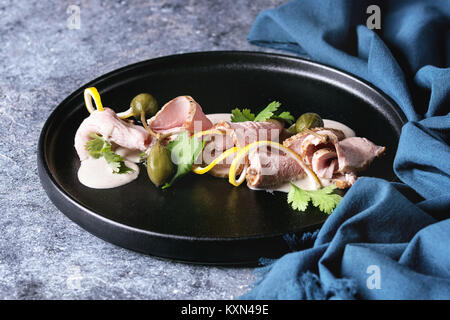 Vitello tonnato italian dish. Thin sliced veal with tuna sauce, capers and coriander served on black plate with blue textile over blue texture backgro Stock Photo