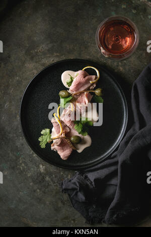 Vitello tonnato italian dish. Thin sliced veal with tuna sauce, capers and coriander served on black plate with glass of rose wine over old dark metal Stock Photo