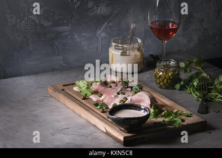 Vitello tonnato italian dish. Thin sliced veal with tuna sauce, capers and coriander served on wooden serving board, glass of rose wine and ingredient Stock Photo
