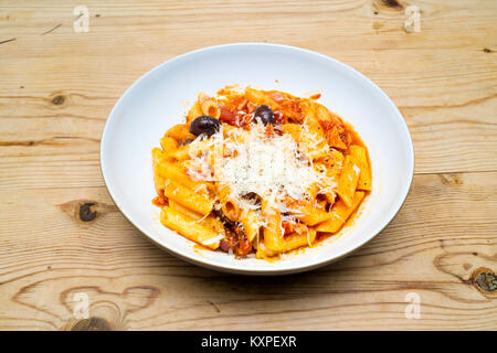 Food Penne Pasta Tonno made with tuna, bacon, tomatoes and Basil, Kalamata Olives, and Pecorino Romano grated cheese in a white bowl on a wooden table Stock Photo