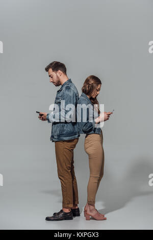 full length side view of young couple in denim jackets standing back to back and using smartphones isolated on grey Stock Photo