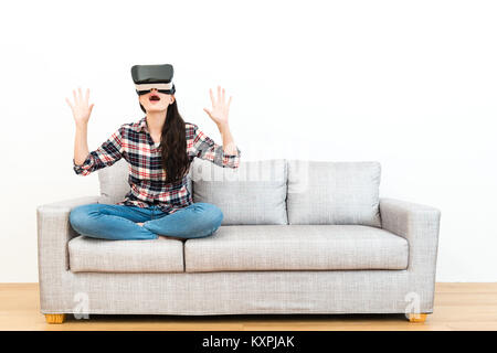 beautiful young woman sitting on wooden floor sofa feeling surprised when she using virtual reality goggles watching 3D simulation video in white back Stock Photo