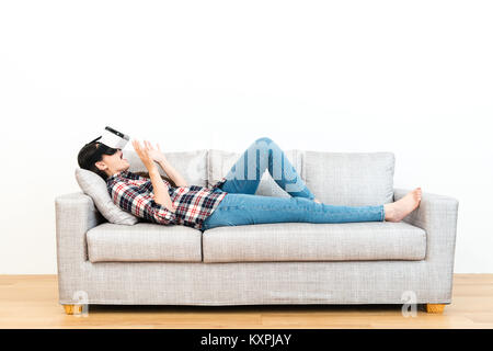 happy cheerful woman lying down on sofa relaxing and using VR technology device watching 3D simulation video feeling surprised. Stock Photo