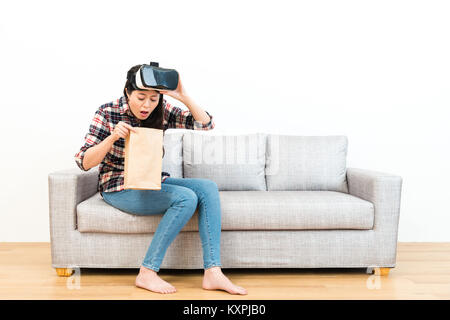 young pretty woman using VR device experiencing 3D game getting dizziness and sitting on wooden floor sofa couch vomiting in white background. Stock Photo