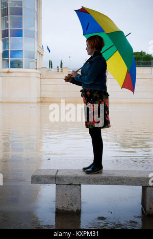a french woman in her thirties stands on a concrete bench with an umbrella by a river which has just burst its banks and created a flood Stock Photo