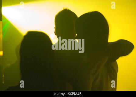 a man hugs his girlfriend next to another female in a nightclub surrounded by glowing yellow disco lights Stock Photo
