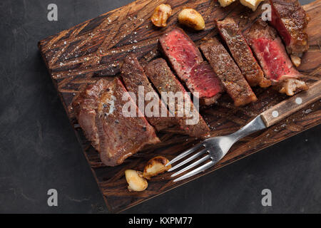 Closeup ready to eat steak new York beef breeds of black Angus with herbs, garlic and butter on a wooden Board. The finished dish for dinner on a dark stone background. Top view Stock Photo