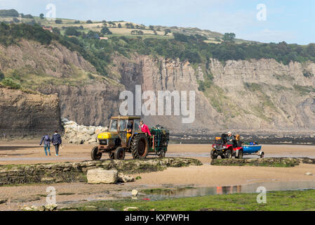 Tractor bringing in Lobster pots ahead of a quad bike and small boat on the beach at Robin Hood's Bay, North Yorkshire, England. Stock Photo