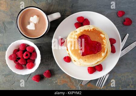 Pancakes with jam in shape of heart, hot chocolate and raspberries over a slate background. Love concept. Stock Photo