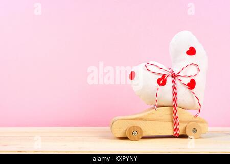 Wooden toy car carrying homemade heart gift against a pink background. Valentines Day or love concept. Stock Photo