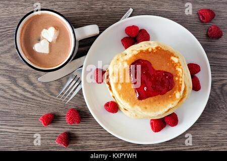 Pancakes with jam in shape of heart, hot chocolate and raspberries over a wood background. Love concept. Stock Photo