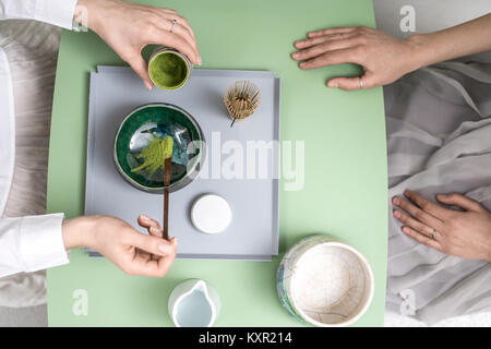Two girls preparing a chinese matcha green tea on the green metal table indoors. One woman puts tea powder into the colorful bowl. On the table there  Stock Photo