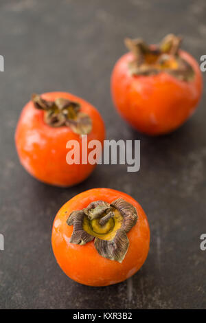 Three persimmons on black rustic tabletop with one persimmon in sharp focus and others blurred. Vertical composition. Shallow depth of field. Stock Photo
