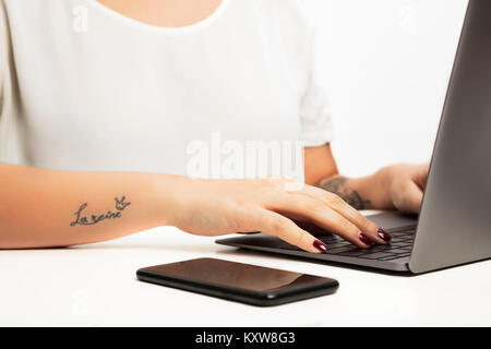 Hands of a young woman typing on a laptop computer in a close up view with a mobile phone alongside. With “la reine” tattoo on arm, meaning “ queen”   Stock Photo
