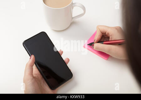 Woman making a sticky note reminder as she copies something off her mobile phone in an over the shoulder view to the blank screen Stock Photo