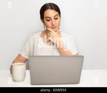 Thoughtful young woman working on a laptop computer looking at the screen with raised eyebrows and a sceptical expression Stock Photo