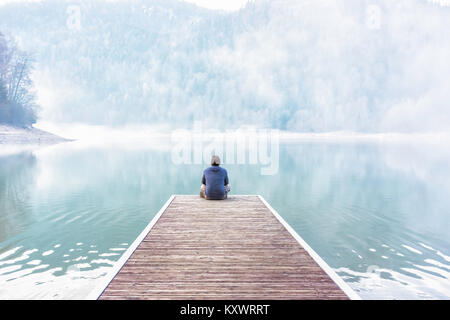Young man sitting and meditating on the dock of a lake on a foggy day of winter, Vosges, France