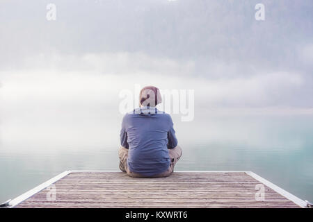 Close-up of a young man sitting and meditating on the dock of a lake on a foggy day of winter, Vosges, France