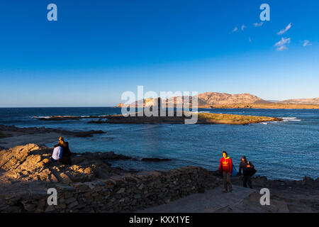 Rear view of people on rocks looking across sea at derelict lighthouse at dusk. La Pelosa, Sardinia. Italy Stock Photo