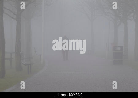 man walking along path towards car headlights on foggy day in the uk Stock Photo