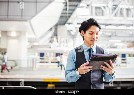 Businessman wearing blue shirt and vest standing on train station platform, holding digital tablet. Stock Photo