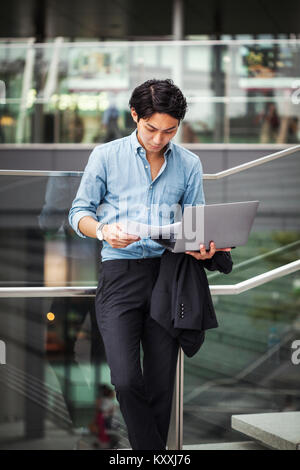 Businessman wearing blue shirt standing outdoors, holding laptop and papers, leaning against glass railing. Stock Photo