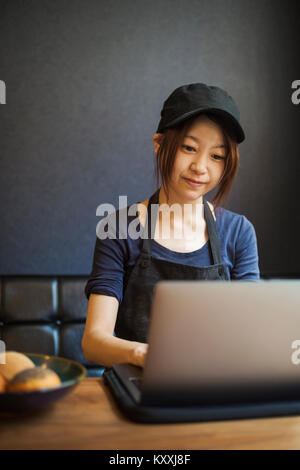 Woman working in a bakery, wearing baseball cap, sitting at table in front of laptop, typing. Stock Photo